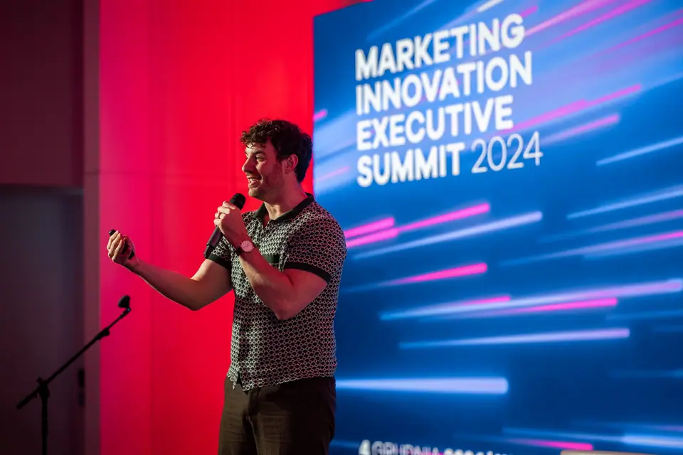 A speaker with a microphone in an energetic gesture in front of the Marketing Innovation Executive Summit 2024 screen